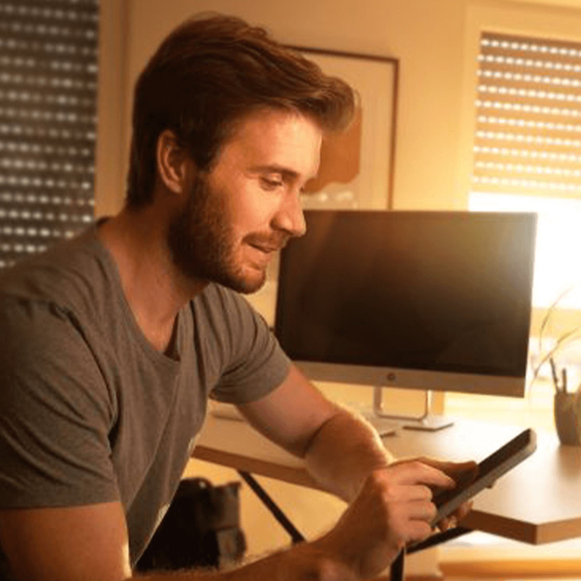 Person sitzt an einem Schreibtisch und benutzt ein Tablet, im Hintergrund ein Monitor