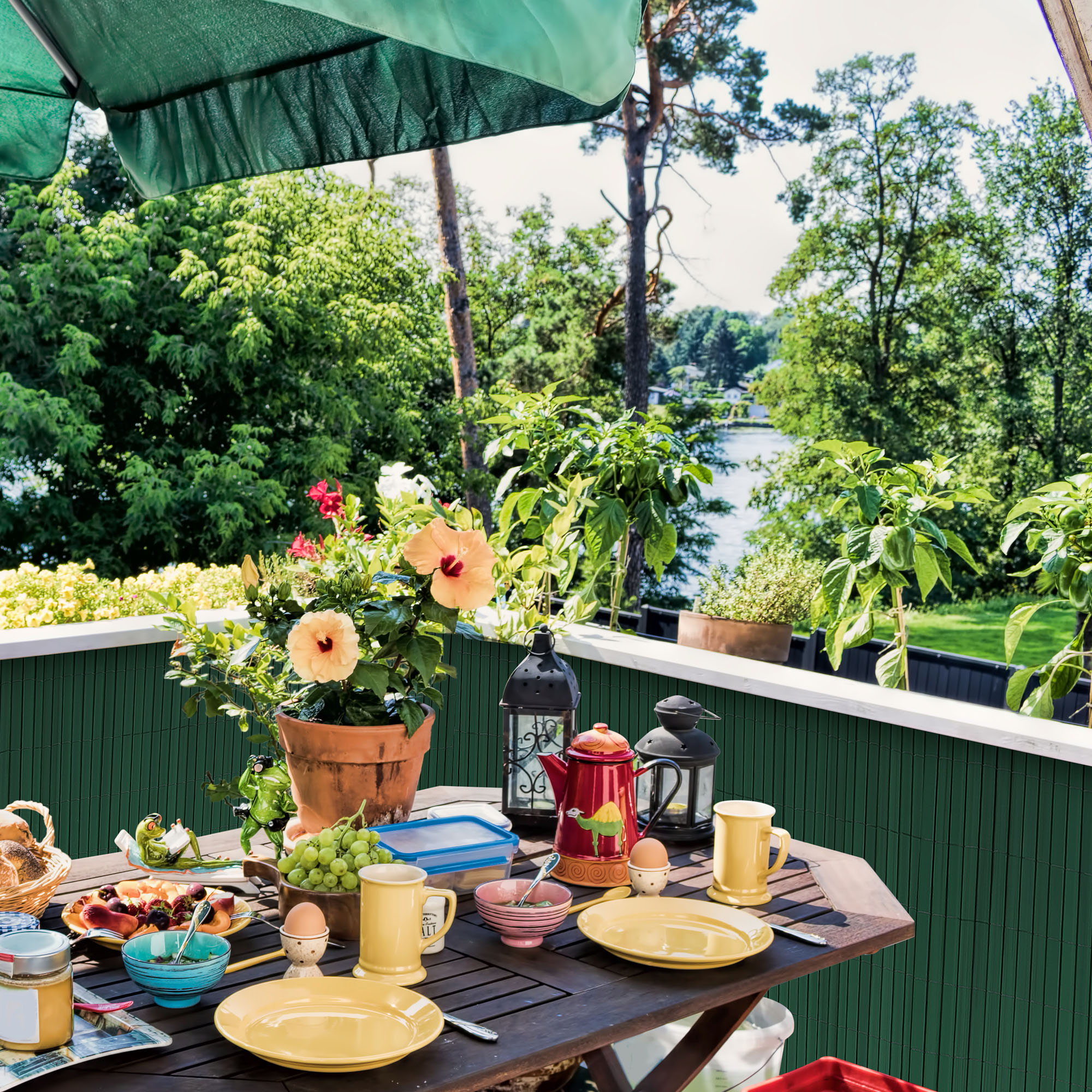 Gedeckter Tisch auf Balkon mit Pflanzen und Sonnenschirm, Frühstücksszene im Freien