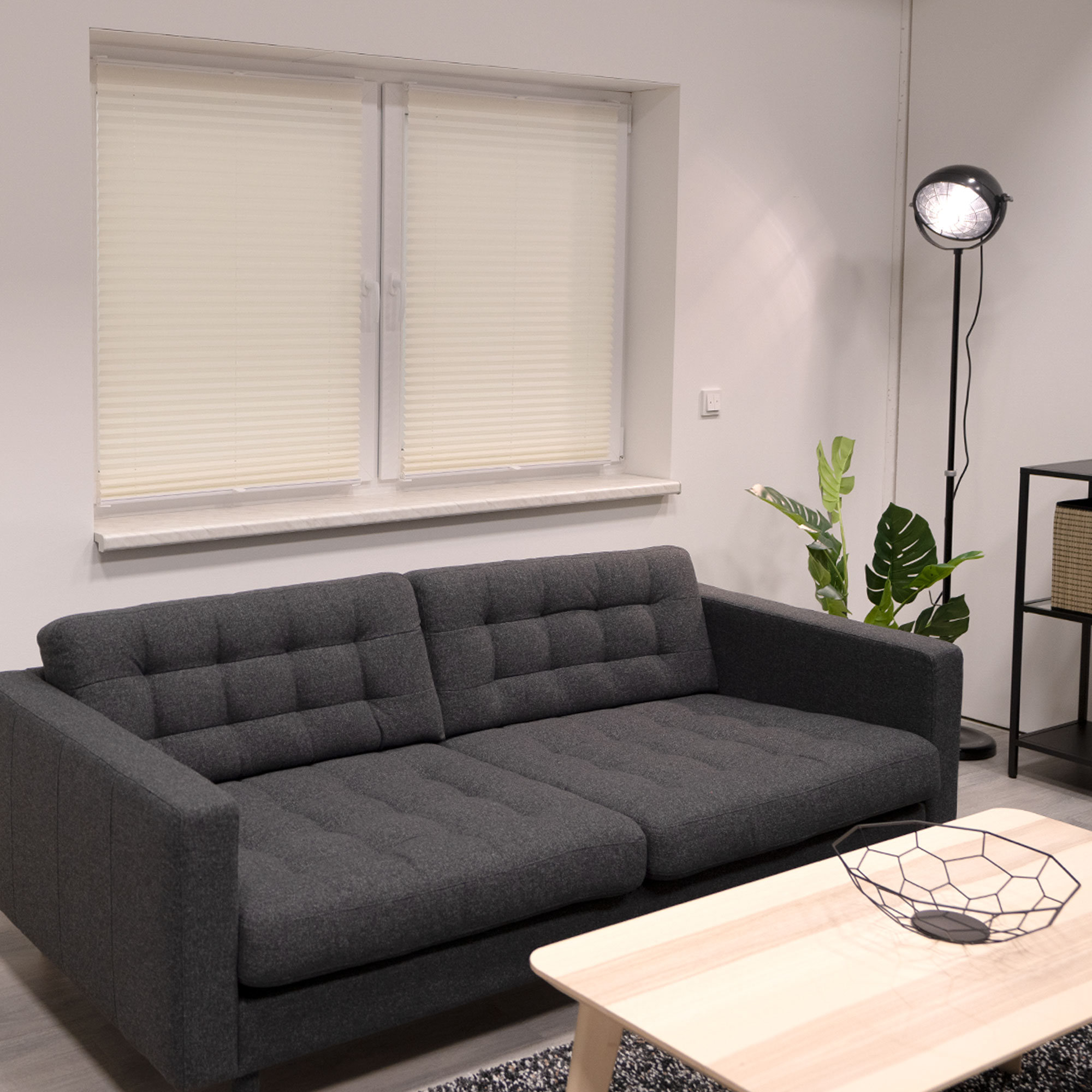 Wohnzimmer mit Sofa, Fenster, Stehlampe, Tisch und Pflanze